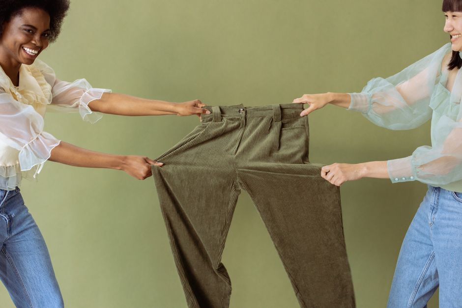 Two women joyfully stretch a pair of green trousers, showcasing fabric flexibility.