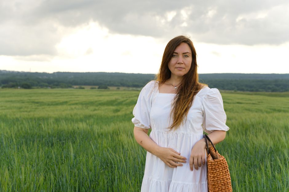 A serene portrait of a woman in a white dress with a woven bag, standing in a lush green field under cloudy skies.