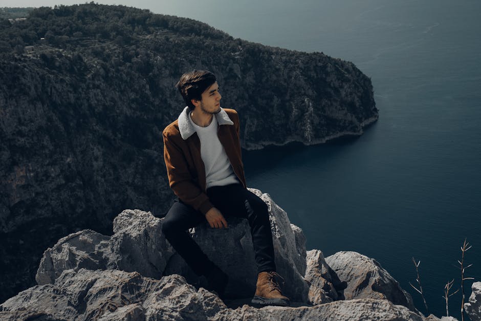 A man in a brown jacket sits on a cliff, gazing at the ocean, surrounded by nature.