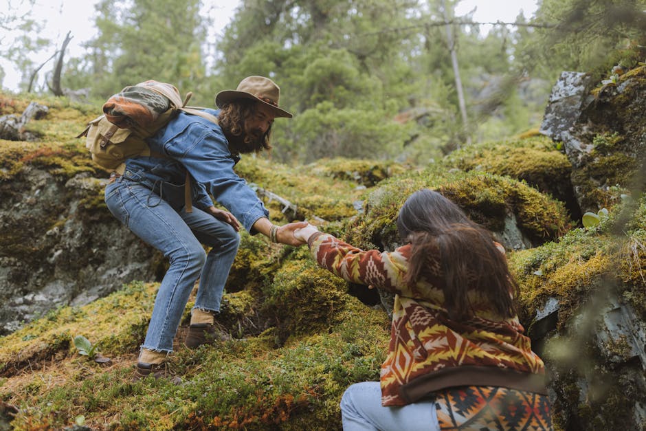 Man helps woman climb a moss-covered rocky slope in a forest setting.