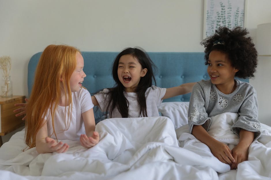 Adorable diverse girls laughing and chatting while sitting on bed under blanket at home