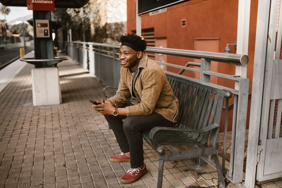 A man sitting on a bench at a city train station while using his phone.