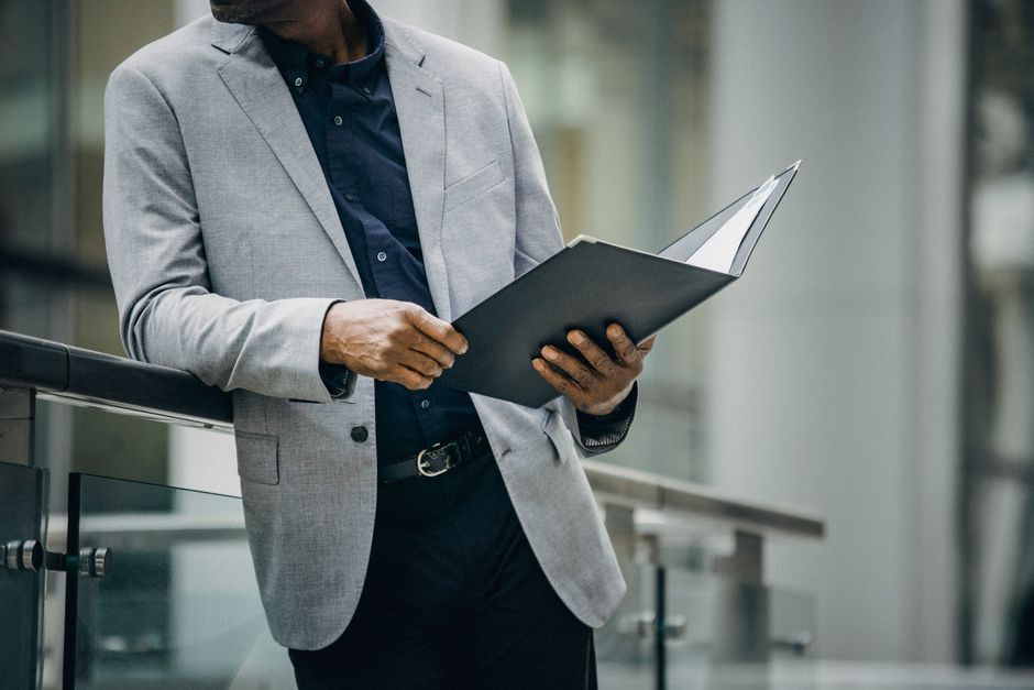 Crop unrecognizable black man in elegant suit reading documents leaning on railing on city street