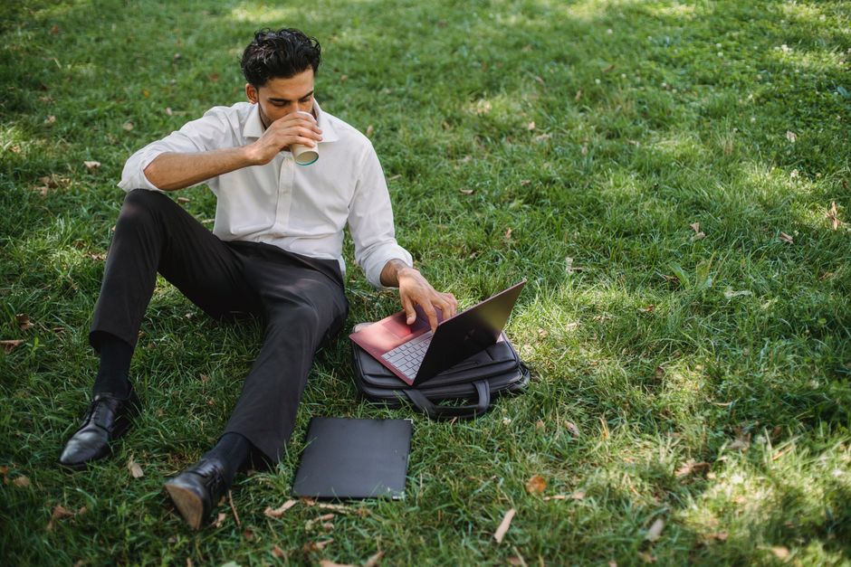 Man in formal attire using laptop and drinking coffee while sitting in a park setting on a sunny day.