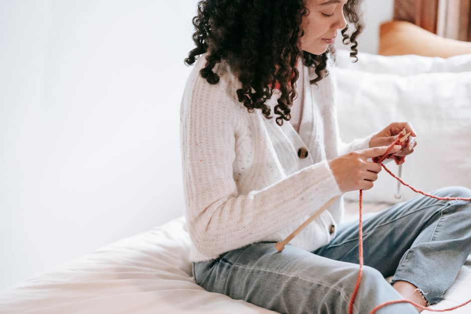 Concentrated young African American female with curly hair wearing white warm cardigan and jeans sitting on bed and knitting with orange threads and crochet