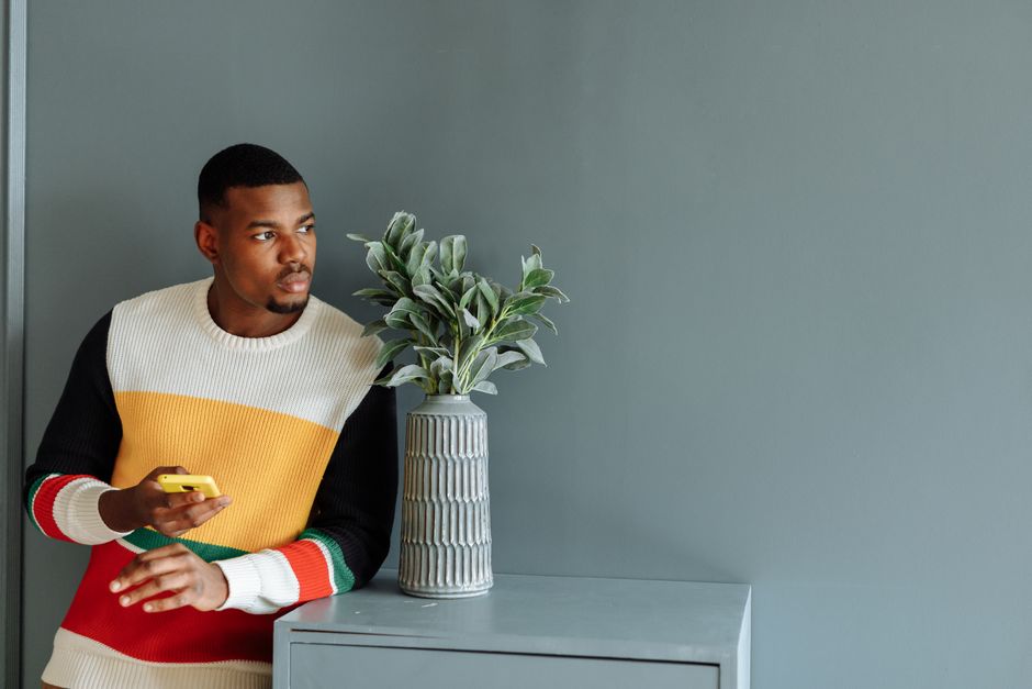A man in a knitted sweater holds a phone, standing by a houseplant on a gray background.