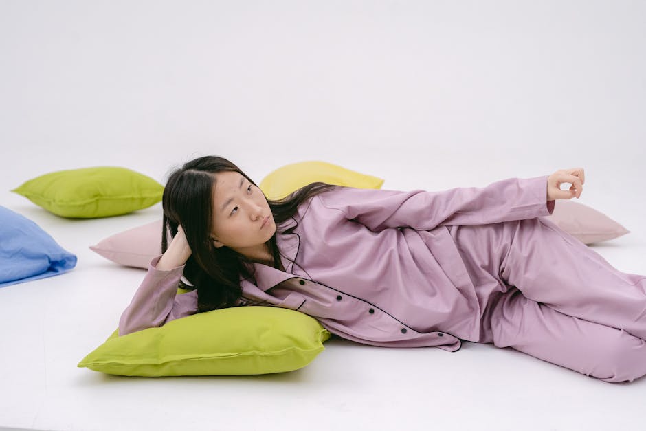 A young woman in pink pajamas lying on colorful pillows, looking thoughtful.