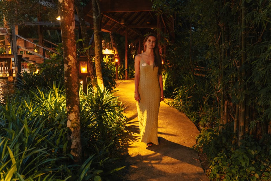 A woman in a yellow dress walks through a tropical garden path, illuminated at night.