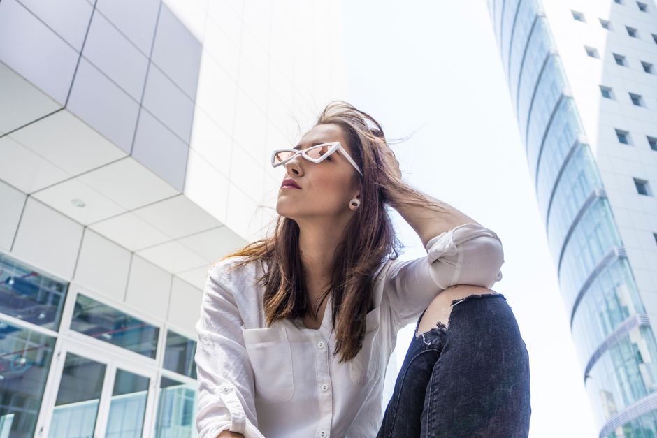 Stylish young woman wearing sunglasses and jeans, posing confidently in modern cityscape.