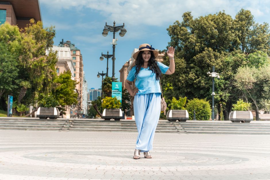 A young woman in a straw hat smiles and waves while exploring Baku's scenic outdoors.