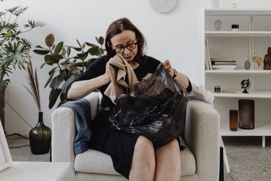 Senior woman seated indoors sorting clothes from a black bag, surrounded by plants and books.