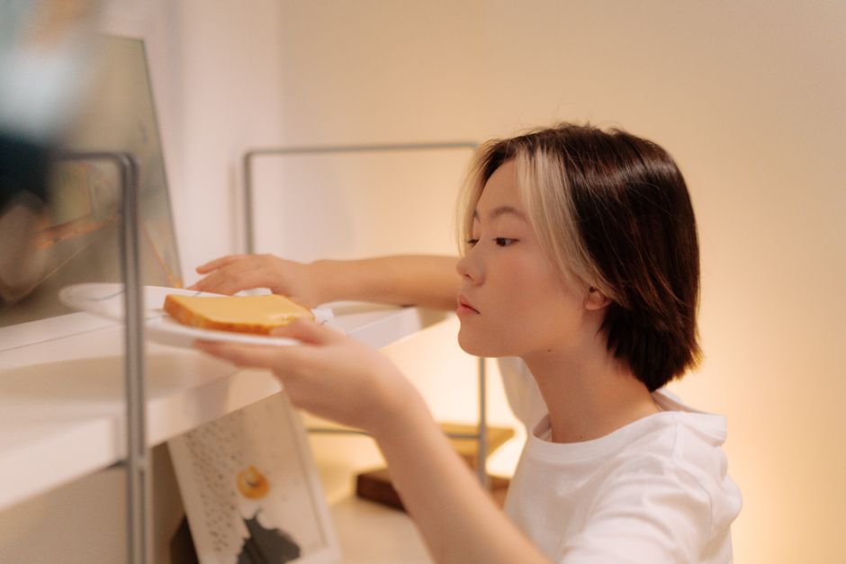 A young Asian woman placing a sandwich on a bookshelf, in a cozy indoor setting.