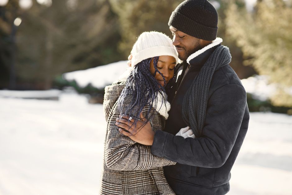 A couple embraces warmly outdoors in winter attire with snow all around, exuding love and happiness.