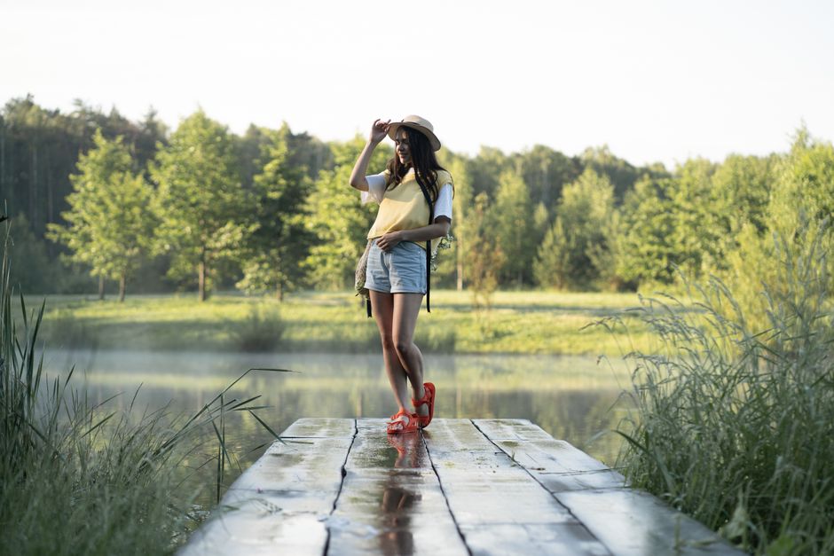 A fashionable woman stands on a lakeside dock surrounded by lush greenery, embodied in a serene summer setting.