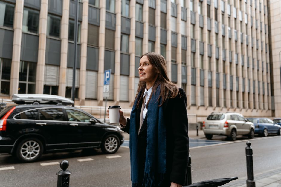 Woman in black coat holding coffee cup, smiling on a city street with buildings and parked cars.