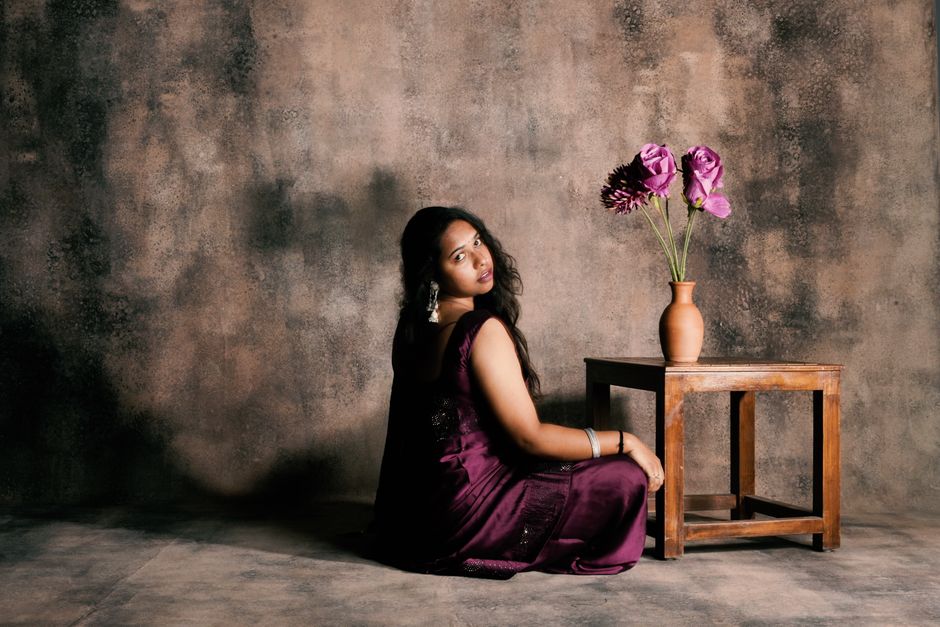 A woman in a purple dress sits gracefully beside a wooden table with a vase of flowers.