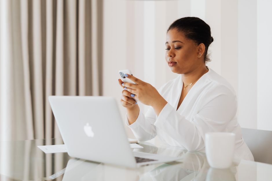A woman in white attire uses her smartphone while working on a laptop at a desk indoors.