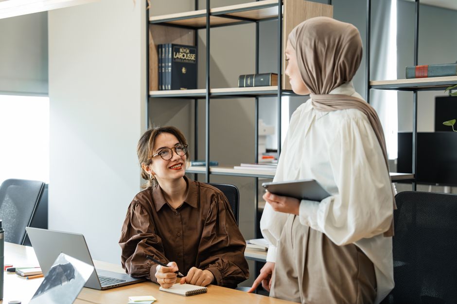 Two women collaborating in a modern office setting, discussing ideas over laptops.
