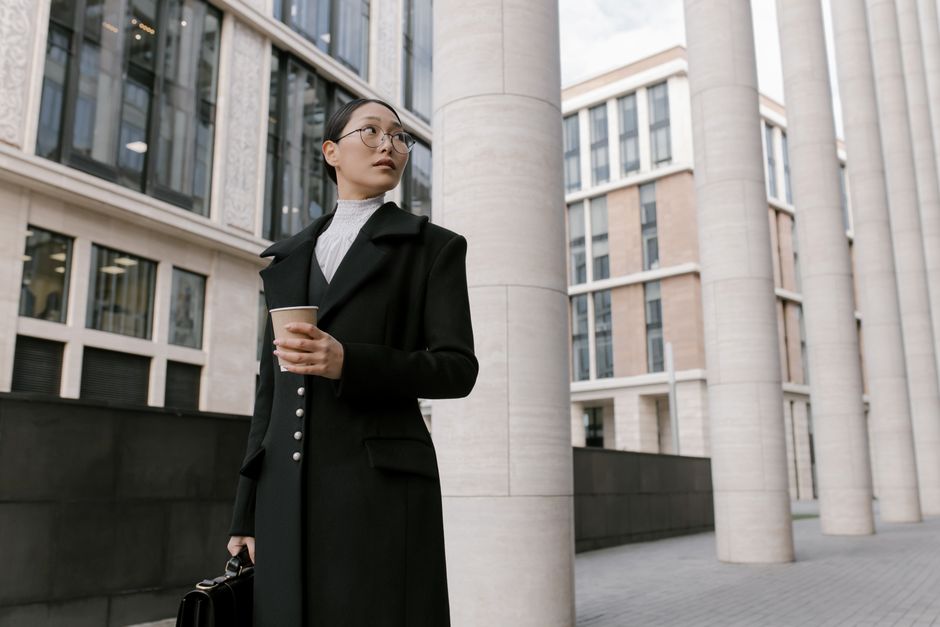 A fashionable businesswoman in a black coat holding a coffee cup outdoors in a corporate setting.