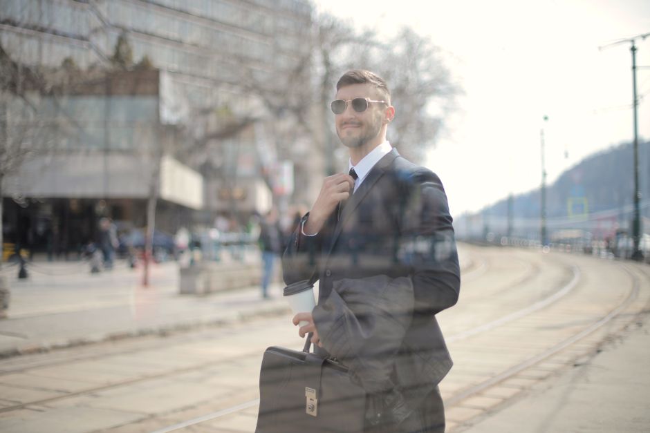 A confident businessman walks in the city, holding a coffee cup and a briefcase, wearing sunglasses.