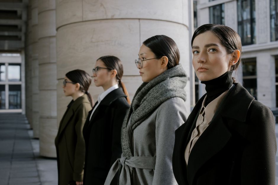 Four professional women in elegant coats walking in an urban setting, showcasing diversity and style.