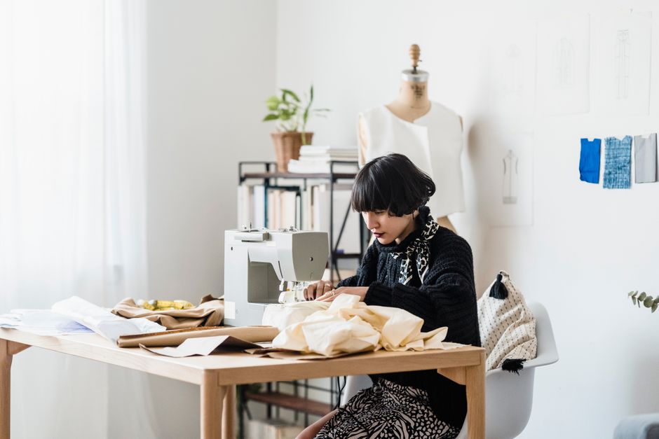 Focused young ethnic female dressmaker with trendy haircut in stylish outfit sewing clothes on machine sitting at wooden table in light atelier