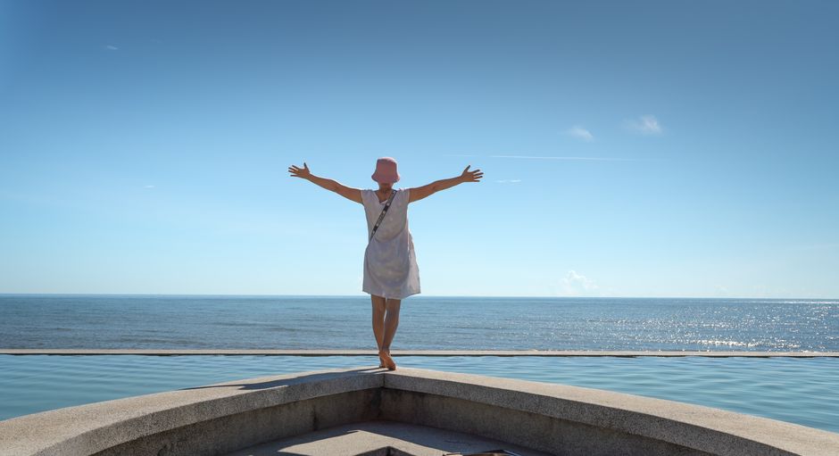 A woman in a sun hat stands by the sea, embracing tranquility under a clear blue sky.