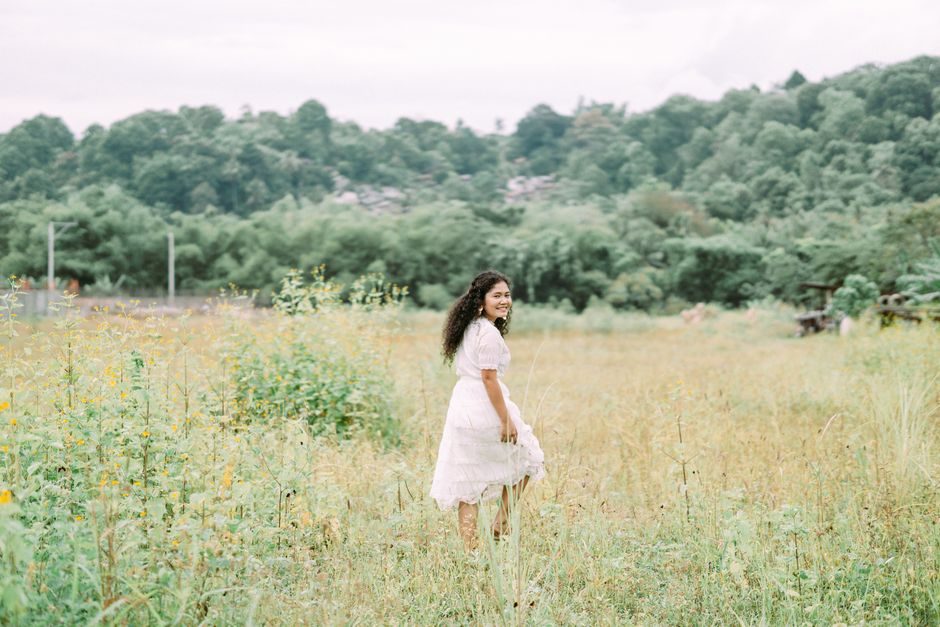 Asian woman in a white dress smiling while walking through a flower-filled field.