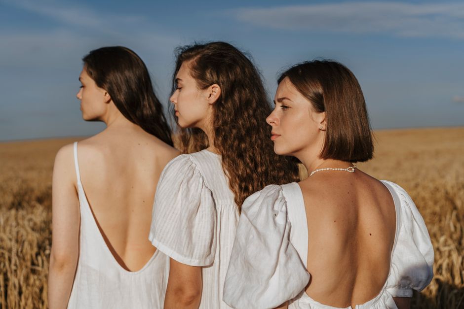 Three women in white dresses stand together in a sunlit wheat field, exuding natural beauty and serenity.
