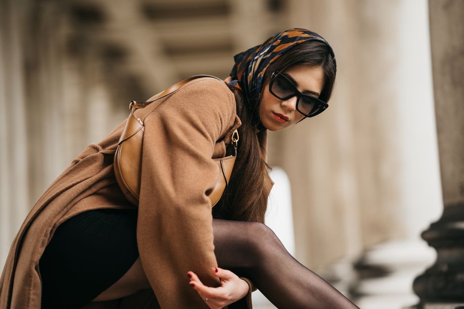 Chic woman in a brown coat with a headscarf poses elegantly in an urban environment.