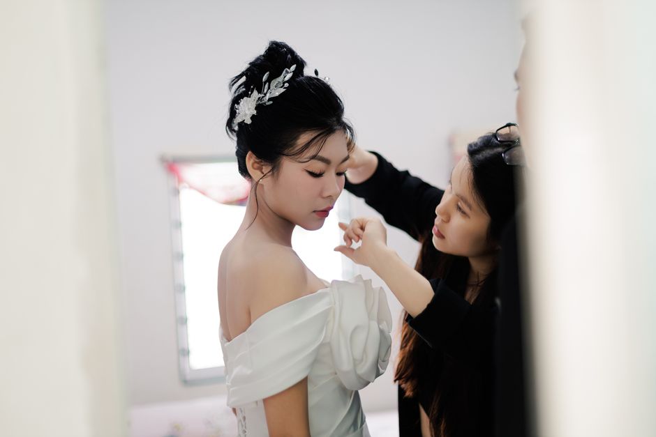 A bride in a white dress being assisted by a makeup artist indoors, showcasing elegance and preparation.