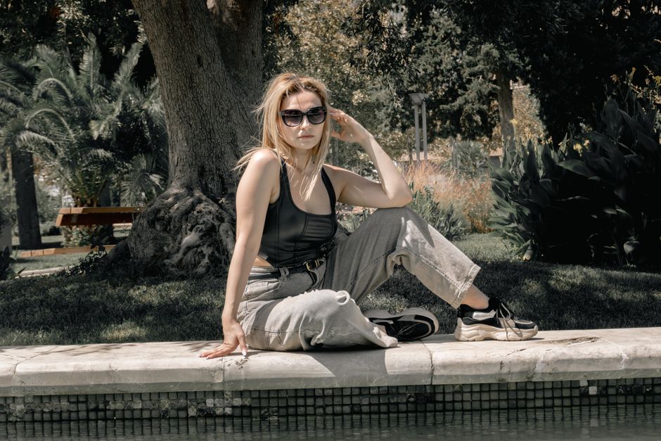 Fashionable woman sitting by a poolside in a garden, wearing sunglasses and casual clothes on a sunny day.