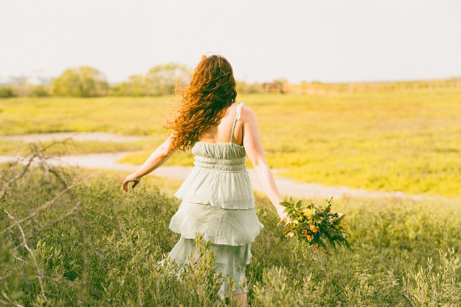 A woman in a light dress walking through a bright countryside meadow holding flowers, capturing a serene and peaceful vibe.
