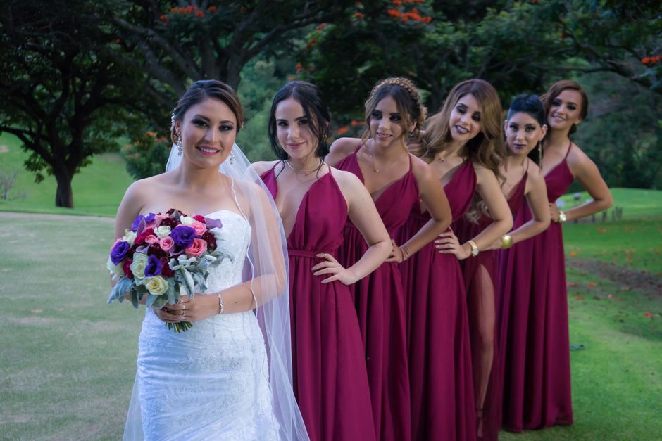 Bride with bridesmaids in burgundy dresses posing outdoors in a lush green setting.