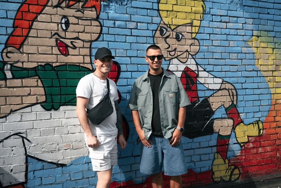 Two men smiling in front of a playful and vibrant street art mural.