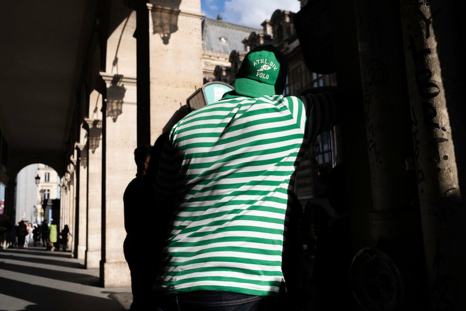 Back view of a man in a green striped shirt standing in sunlight in a Paris colonnade.