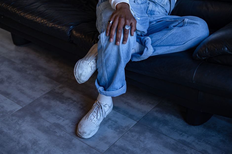 Close-up of a person in denim pants sitting on a sofa, highlighting casual fashion with white sneakers and rings.