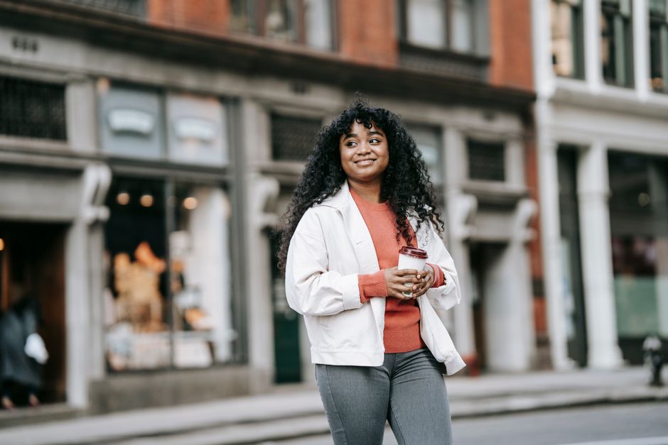 Smiling adult African American female in casual clothes with coffee to go walking on city street and looking away contentedly
