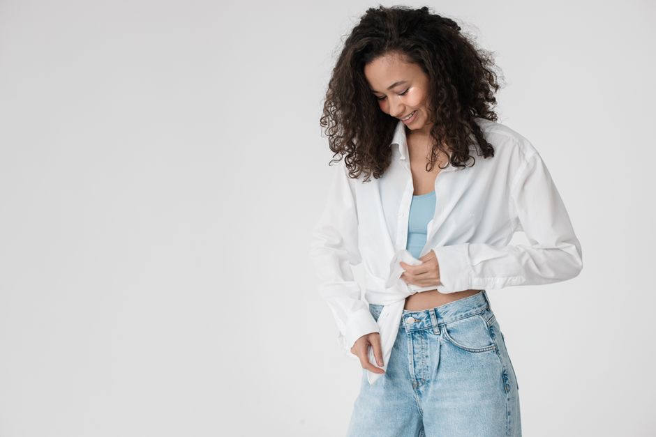 Young woman in a casual outfit smiling and adjusting her denim jeans against a neutral background.